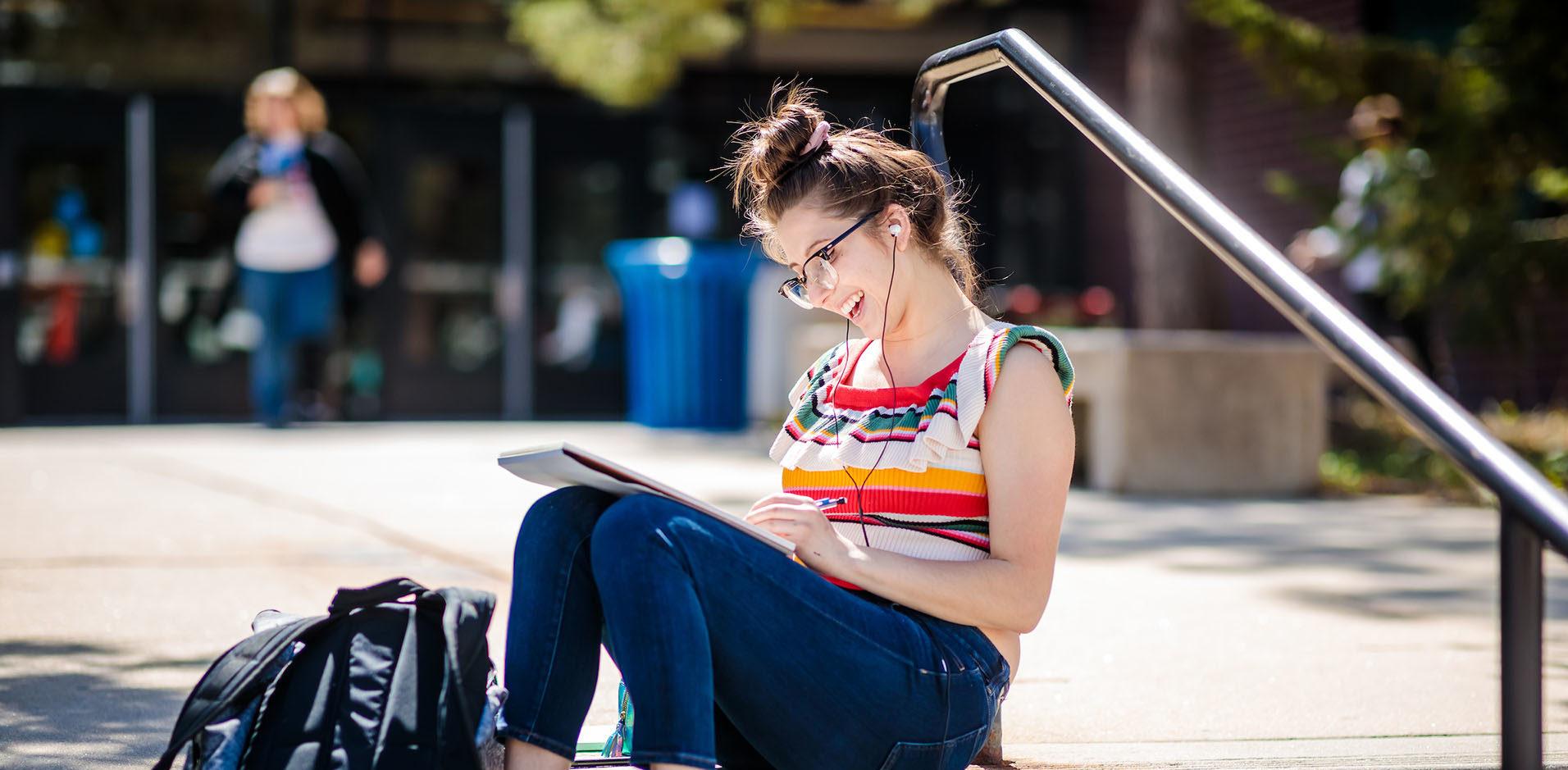 Student sitting on stairs writing in notebook and with headphones