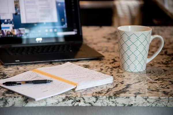 A laptop, notebook and a cup of coffee on a kitchen counter.