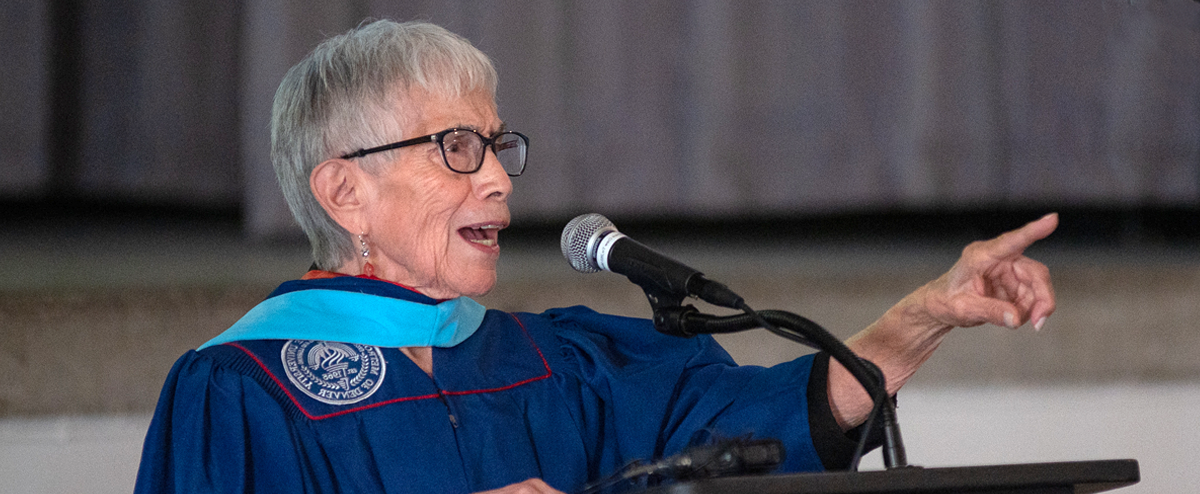 Anna Jo Garcia Haynes wearing MSU Denver regalia, speaking at a lectern with a microphone at the Master of Arts in Teaching Hooding Ceremony, May, 2023.