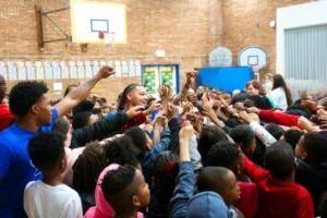 图为一群先生和年轻学生站在学校体育馆里，举起双手，指着中心.