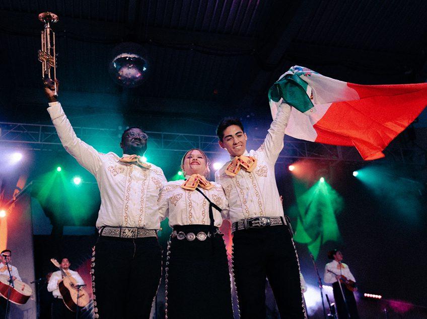 three mariachi performers waving to audience with trumpet and Mexican flag